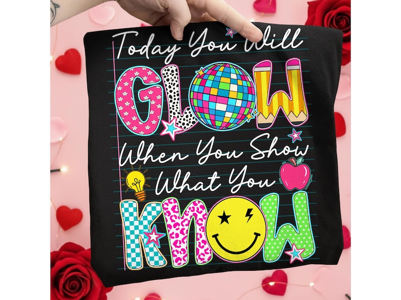 May include: Black t-shirt with the words "Today You Will GLOW When You Show What You KNOW" in colorful, playful lettering. The design includes a disco ball, pencils, a lightbulb, an apple, and a smiley face.