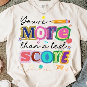 May include: Cream-colored t-shirt with the text "You're More Than a Test Score" in colorful, block letters. The design includes a pencil, stars, an apple, and other school-themed graphics.
