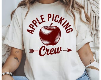 May include: Cream-colored t-shirt with the words "APPLE PICKING CREW" in burgundy, featuring a detailed apple graphic. An arrow points to the right, below the word "Crew". The shirt is a casual, comfortable style.