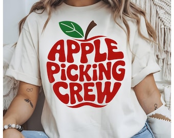 May include: Cream-colored t-shirt featuring a large red apple graphic with the words "APPLE PICKING CREW" in a bold, retro font. The apple has a green leaf and brown stem. The shirt is casual and suitable for fall activities.