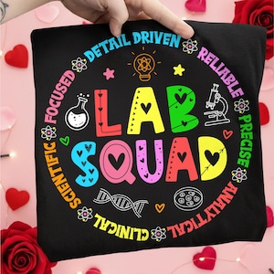 Puede incluir: Camiseta negra con las palabras "LAB SQUAD" en letras coloridas, rodeada de iconos científicos y frases como "Detail Driven" y "Scientific". El diseño incluye corazones, una bombilla y un microscopio.
