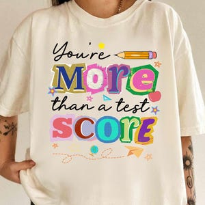 May include: Cream-colored t-shirt with the text "You're More than a test SCORE" in colorful, playful lettering. A pencil graphic and various decorative elements like stars and a paper airplane are included in the design. The shirt is casual wear.