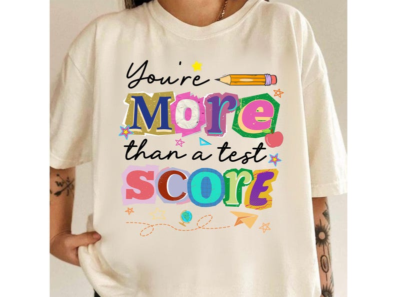 May include: Cream-colored t-shirt with the text "You're More than a test SCORE" in colorful, playful lettering. A pencil graphic and various decorative elements like stars and a paper airplane are included in the design. The shirt is casual wear.