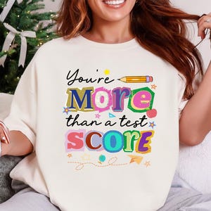 May include: Cream-colored sweatshirt with the text "You're More than a test SCORE" in colorful, playful lettering. The design includes a pencil, stars, and other decorative elements, perfect for teachers or students.