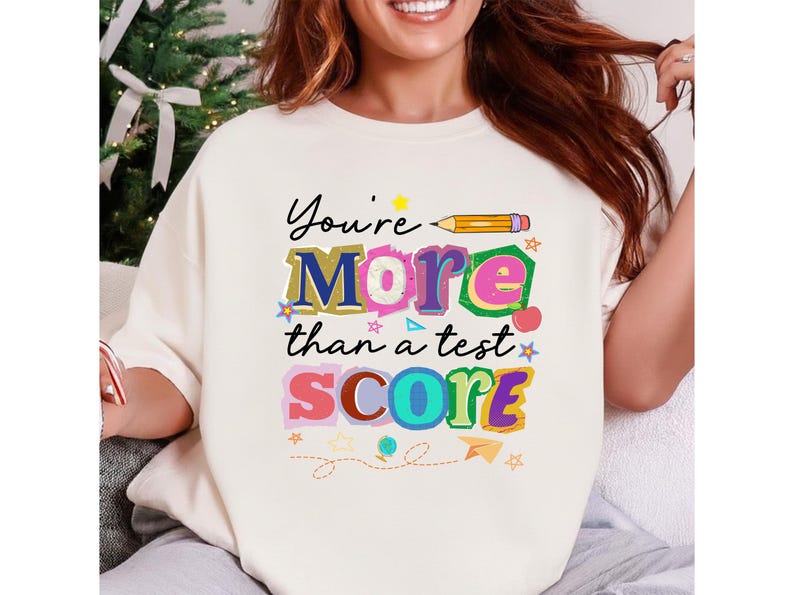 May include: Cream-colored sweatshirt with the text "You're More than a test SCORE" in colorful, playful lettering. The design includes a pencil, stars, and other decorative elements, perfect for teachers or students.