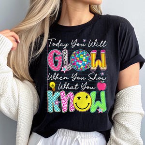 May include: Black t-shirt with the text "Today You Will GLOW When You Show What You KNOW." The word "GLOW" has a disco ball, star, and pencil design. The word "KNOW" has a smiley face and star design. The shirt is a ribbed knit.