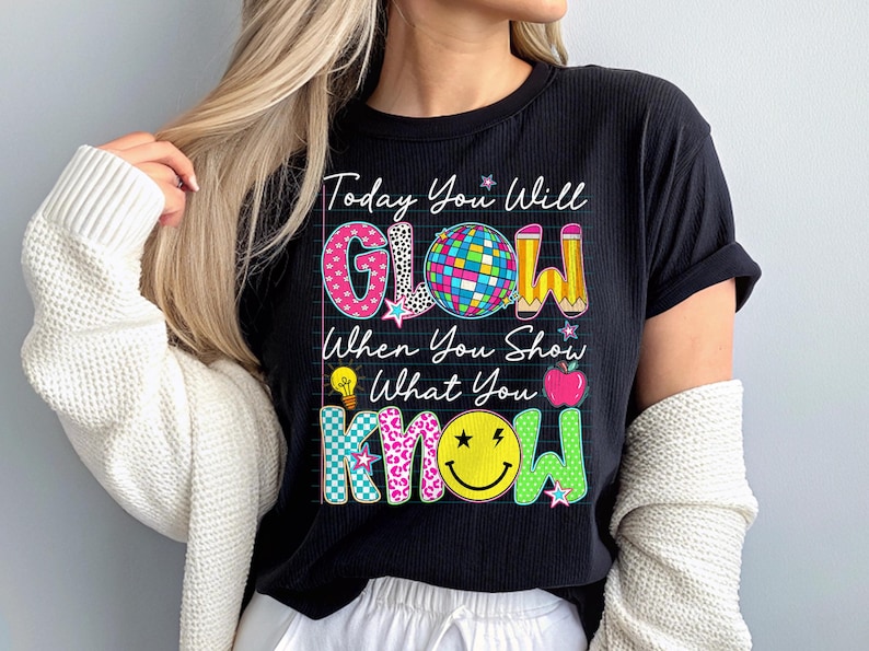 May include: Black t-shirt with the text "Today You Will GLOW When You Show What You KNOW." The word "GLOW" has a disco ball, star, and pencil design. The word "KNOW" has a smiley face and star design. The shirt is a ribbed knit.