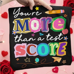 May include: Black t-shirt with the text "You're More than a test SCORE" in colorful, layered letters. A pencil graphic and various stars, a globe, and a paper airplane are also featured.