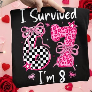 Może przedstawiać: Czarna koszulka z napisem "I Survived I'm 8". Numer 6 jest w szachownicę, a numer 7 ma różowy nadruk w panterkę. Oba numery mają różowe kokardy i gwiazdki. Serca są rozrzucone wokół wzoru.