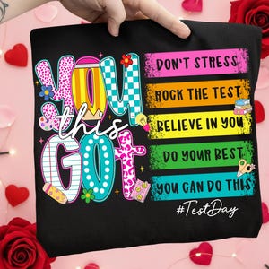 May include: Black t-shirt with the words "YOU GOT THIS" in colorful, playful lettering. The shirt also features the phrases "DON'T STRESS, ROCK THE TEST, BELIEVE IN YOU, DO YOUR BEST, YOU CAN DO THIS" and the hashtag #TestDay.