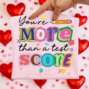 May include: A light pink t-shirt with the text "You're More than a test SCORE" in colorful, layered letters. A hand points to the shirt, surrounded by red heart-shaped balloons and pink polka dots.