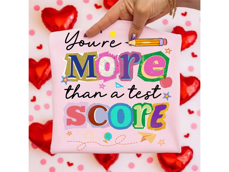 May include: A light pink t-shirt with the text "You're More than a test SCORE" in colorful, layered letters. A hand points to the shirt, surrounded by red heart-shaped balloons and pink polka dots.