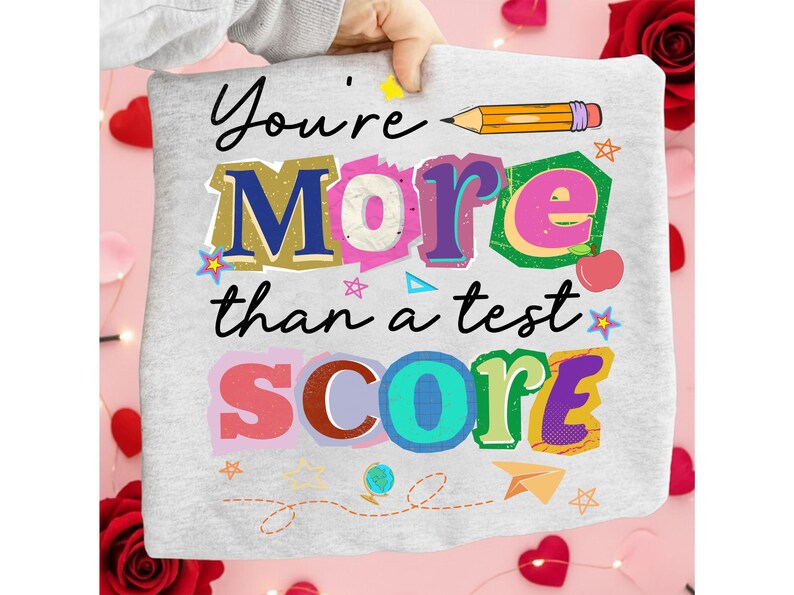 May include: A light gray t-shirt with the text "You're More than a test SCORE" in colorful, layered letters. The design includes a pencil, apple, stars, and other school-themed graphics. The shirt is held up by a hand.