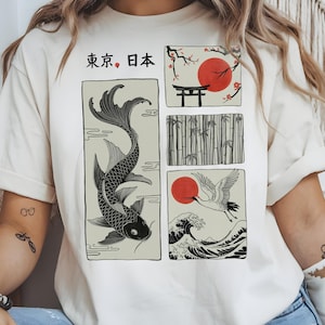 Puede incluir: Camiseta color crema con ilustraciones de inspiración japonesa. El diseño incluye una carpa koi, un sol rojo, una grulla, bambú y una puerta tradicional. El texto japonés "Tokyo, Japón" está impreso en la parte superior. La camiseta tiene mangas enrolladas.