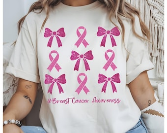 May include: Cream-colored t-shirt featuring a design of pink ribbons and bows, with a glittery texture. The phrase "#Breast Cancer Awareness" is printed in a cursive font below the design. The shirt is a casual style.