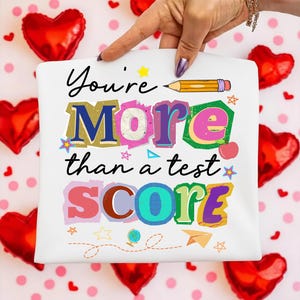 May include: White square with the text "You're More than a test SCORE" in colorful, playful lettering. A yellow pencil graphic is above the word "More". The background includes red heart-shaped balloons and pink polka dots.