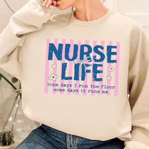 May include: A cream-colored sweatshirt with a graphic that reads "NURSE LIFE" in blue, with pink vertical stripes in the background. The text "some days I run the floor some days it runs me" is below the main text. There are heart and flower graphics.