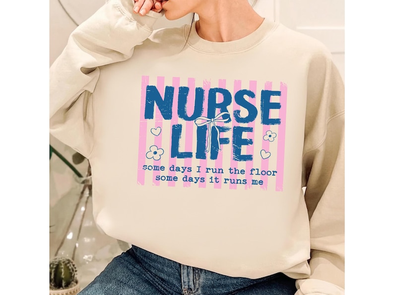 May include: A cream-colored sweatshirt with a graphic that reads "NURSE LIFE" in blue, with pink vertical stripes in the background. The text "some days I run the floor some days it runs me" is below the main text. There are heart and flower graphics.