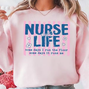 May include: A light pink sweatshirt with the words "NURSE LIFE" in blue block letters. The text is set against a pink and white striped background. The phrase "some days I run the floor some days it runs me" is below.