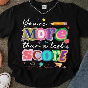 May include: Black t-shirt with the text "You're More than a test SCORE" in colorful, playful lettering. The design includes a pencil, stars, an apple, and a paper airplane, perfect for teachers or students.
