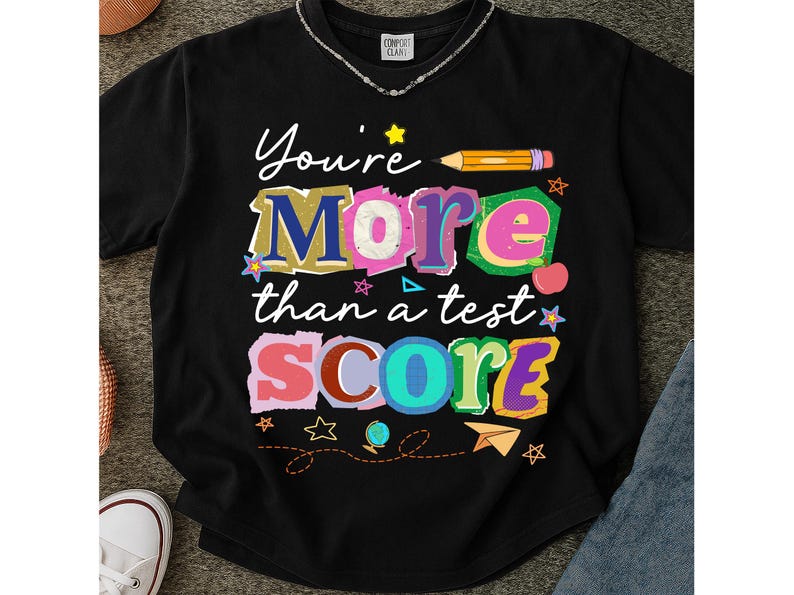 May include: Black t-shirt with the text "You're More than a test SCORE" in colorful, playful lettering. The design includes a pencil, stars, an apple, and a paper airplane, perfect for teachers or students.