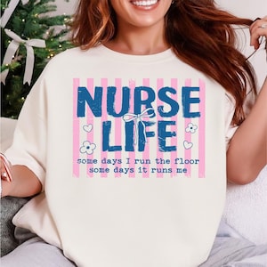May include: Cream-colored sweatshirt with the words "NURSE LIFE" in blue block letters, set against pink striped background. The text "some days I run the floor some days it runs me" is below. Floral and heart graphics are also present.