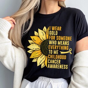May include: Black t-shirt with a sunflower graphic and the text "I Wear Gold For Someone Who Means Everything To Me Childhood Cancer Awareness." The sunflower is yellow and black. The shirt is ribbed.