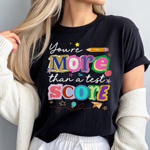 May include: Black t-shirt with the text "You're More than a test SCORE" in colorful, playful lettering. The design includes stars, a pencil, and a paper airplane. The shirt is ribbed and has rolled-up sleeves.