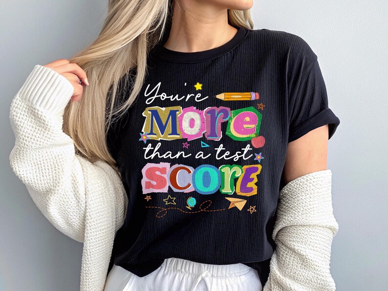 May include: Black t-shirt with the text "You're More than a test SCORE" in colorful, playful lettering. The design includes stars, a pencil, and a paper airplane. The shirt is ribbed and has rolled-up sleeves.