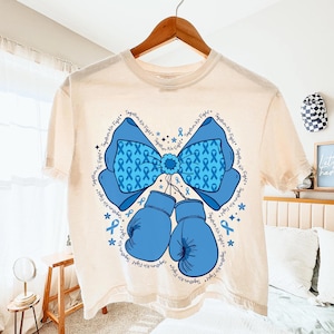 May include: Cream-colored t-shirt featuring a blue bow and boxing gloves design. The bow is filled with blue ribbons and surrounded by the text "Together We Fight." Blue boxing gloves hang below the bow, with stars and ribbons scattered around.