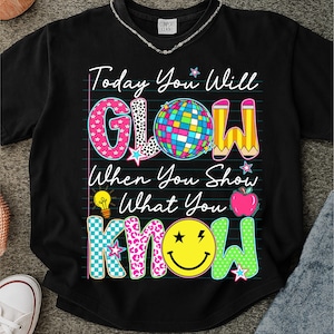 May include: Black t-shirt with the text "Today You Will GLOW When You Show What You KNOW." The word "GLOW" is in pink, black and white, and blue. The word "KNOW" is in teal, yellow, and green. The shirt has a disco ball, pencil, lightbulb, and apple graphic.