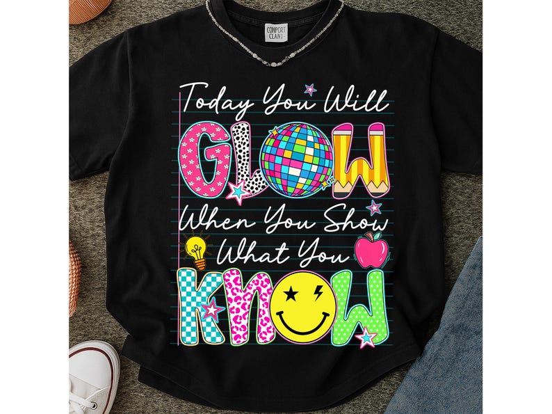 May include: Black t-shirt with the text "Today You Will GLOW When You Show What You KNOW." The word "GLOW" is in pink, black and white, and blue. The word "KNOW" is in teal, yellow, and green. The shirt has a disco ball, pencil, lightbulb, and apple graphic.