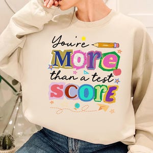 May include: Cream-colored sweatshirt with the text "You're More than a test SCORE" in colorful, playful lettering. A pencil and various decorative elements accompany the text, creating a cheerful and encouraging design.