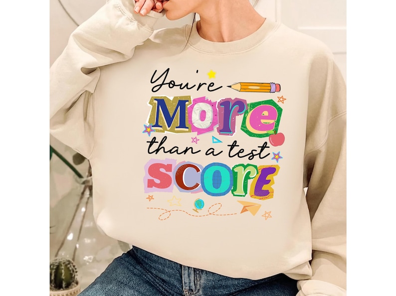 May include: Cream-colored sweatshirt with the text "You're More than a test SCORE" in colorful, playful lettering. A pencil and various decorative elements accompany the text, creating a cheerful and encouraging design.
