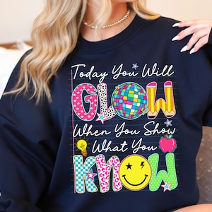 May include: Navy blue sweatshirt with the text "Today You Will GLOW When You Show What You KNOW." The design includes a disco ball, pencils, a lightbulb, an apple, and a smiley face.