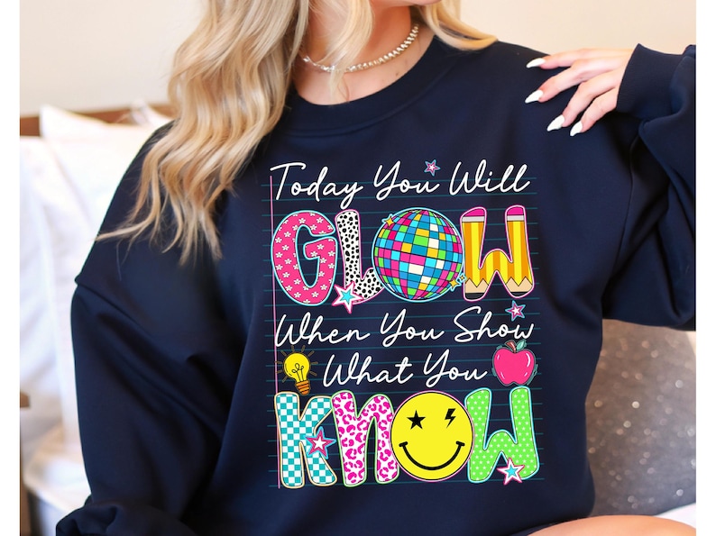 May include: Navy blue sweatshirt with the text "Today You Will GLOW When You Show What You KNOW." The design includes a disco ball, pencils, a lightbulb, an apple, and a smiley face.