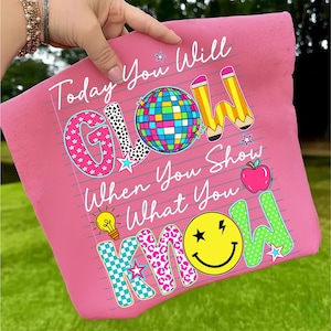 May include: Pink pillow with the text "Today You Will GLOW When You Show What You KNOW." The word "GLOW" has a disco ball design. The word "KNOW" has a smiley face design. Other graphics include pencils, stars, an apple, and a lightbulb.