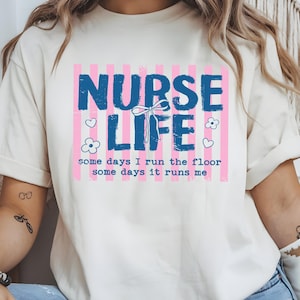 May include: Cream-colored t-shirt with a graphic that reads "NURSE LIFE" in blue, set against a pink striped background. The design includes hearts, flowers, and the text "some days I run the floor, some days it runs me."