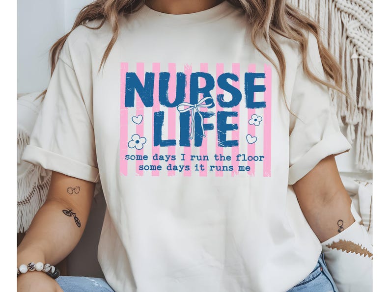 May include: Cream-colored t-shirt with a graphic that reads "NURSE LIFE" in blue, set against a pink striped background. The design includes hearts, flowers, and the text "some days I run the floor, some days it runs me."