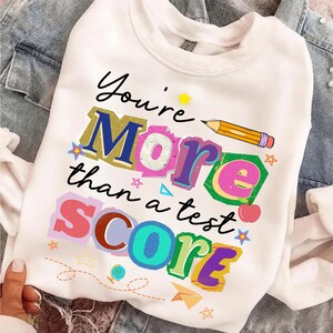 May include: White sweatshirt with the text "You're More than a test SCORE" in colorful, layered letters. A pencil graphic is above the word "More". The design includes stars, a paper airplane, and a small apple.