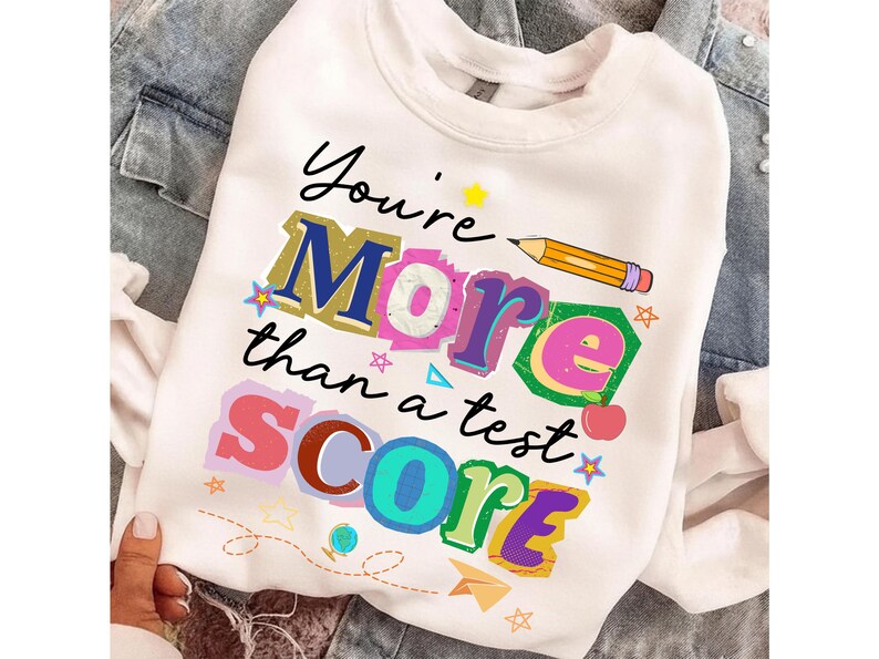 May include: White sweatshirt with the text "You're More than a test SCORE" in colorful, layered letters. A pencil graphic is above the word "More". The design includes stars, a paper airplane, and a small apple.