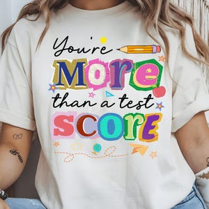 May include: Cream-colored t-shirt with the message "You're More than a test SCORE" in colorful, playful lettering. A yellow pencil graphic and various small illustrations like stars and an apple decorate the design. The shirt is casual and suitable for everyday wear.