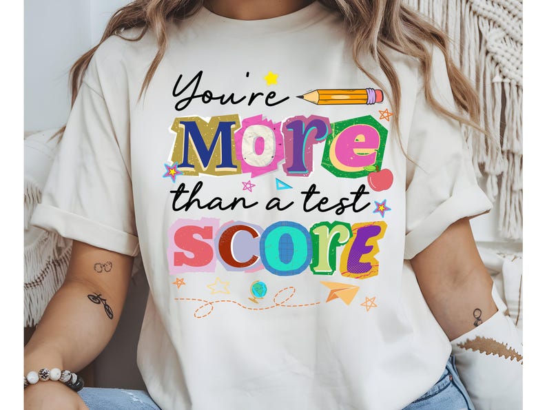 May include: Cream-colored t-shirt with the message "You're More than a test SCORE" in colorful, playful lettering. A yellow pencil graphic and various small illustrations like stars and an apple decorate the design. The shirt is casual and suitable for everyday wear.