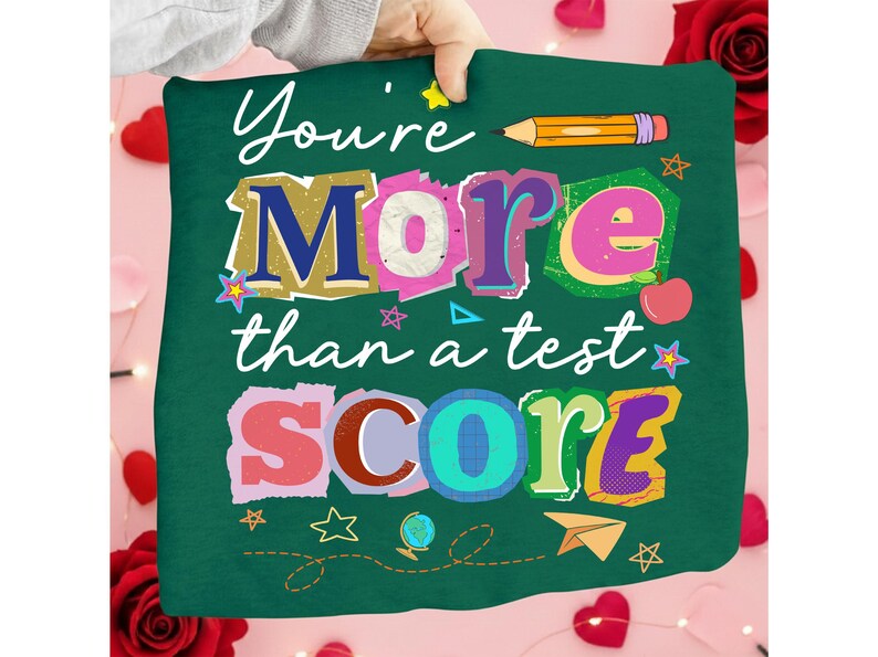 May include: A green pillow with the words "You're More than a test SCORE" in colorful, layered text. The design includes a pencil, stars, an apple, a globe, and a paper airplane. The pillow is held by a person.