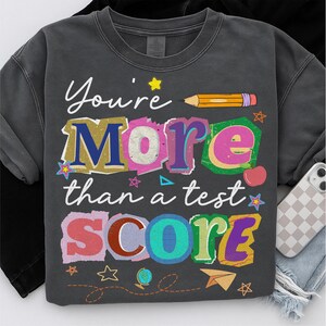 May include: Dark gray sweatshirt with the text "You're More than a test SCORE" in colorful, layered lettering. A yellow pencil, stars, and other school-themed graphics are included in the design.