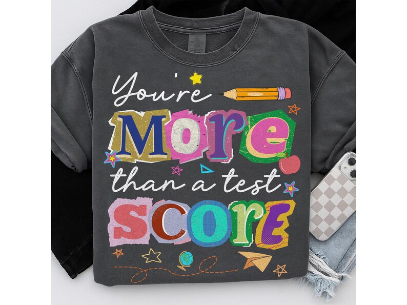May include: Dark gray sweatshirt with the text "You're More than a test SCORE" in colorful, layered lettering. A yellow pencil, stars, and other school-themed graphics are included in the design.
