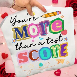 May include: White pillowcase with the text "You're More than a test SCORE" in colorful, block letters. A yellow pencil and various small illustrations, including stars and an apple, are also featured.