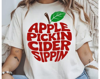May include: Cream-colored t-shirt featuring a large red apple design with the words "APPLE PICKIN CIDER SIPPIN" in a bold, retro font. The apple has a brown stem and a green leaf. A casual, fall-themed graphic tee.
