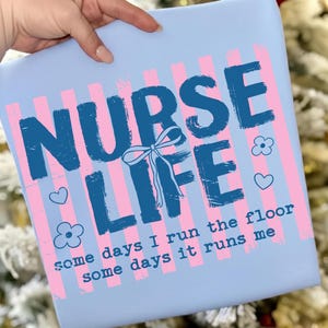 May include: A light blue square with the words "NURSE LIFE" in dark blue, with a bow, flowers, and hearts. Pink and white vertical stripes are behind the text. The text "some days I run the floor some days it runs me" is at the bottom.