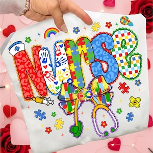 May include: White t-shirt with the word "NURSE" in colorful, patterned letters. The design includes a rainbow, puzzle pieces, a stethoscope, and a syringe. The shirt is held up by a hand.